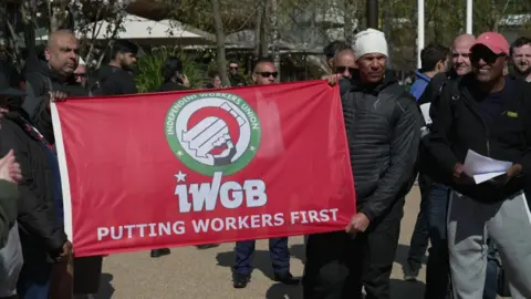 Protesters gather outdoors holding a red banner from the Independent Workers Union of Great Britain (IWGB) that reads "Putting Workers First," with several people standing behind.
