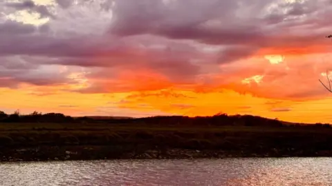 Weather Watcher/Space Walker A stunning photo at dusk with a river in the foreground and the bank behind, and the sky is a flaming yellowy-orange colour, with ominous grey clouds tinged with the sun's rays.