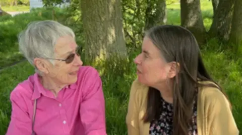 Nikki Lushington Two women facing each other and sitting among trees in dappled sunlight.