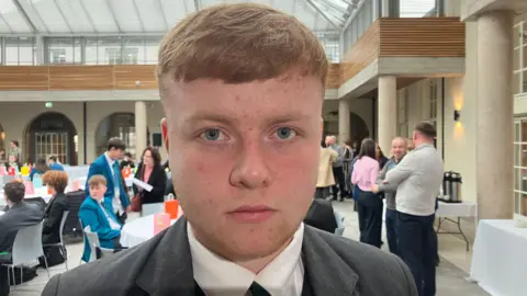 A teenage boy, wearing a white shirt and grey blazer. He has short red hair. 