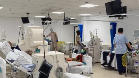 The Renal Department at Sunderland Royal Hospital. Three medical practioners tend to patients on dialysis, they are dressed in medical scrubs, while dialysis machines can be seen near the patients beds. 