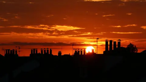 Marie-Pierre Garroway Sunset silhouette of rooftops and chimneys against a vibrant orange and yellow sky, with scattered clouds enhancing the scene.