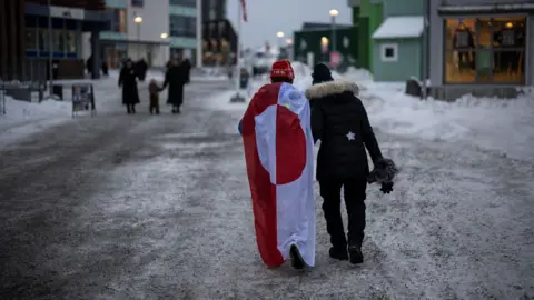 Two people walk down an icy street. One is draped in a Greenland flag.