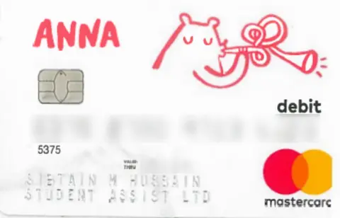 NCA A debit card with a predominantly white background and colourful decorative elements. Near the top left, the brand “ANNA” appears in bright pink, handwritten-style text. To the right side of the card, there is a playful illustration of a small bear-like character blowing a trumpet, drawn in pink line art. The word “debit” is printed in lowercase black letters on the right side. Toward the bottom right corner, the Mastercard logo is displayed, featuring its two overlapping red and yellow circles. A chip is visible on the left side of the card. The lower portion contains embossed card details, including a card number and the cardholder’s name.