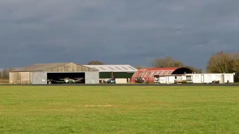 Evelyn Simak/Geograph Hangars with small planes located inside can be seen in the distance, in front of a grass surface.