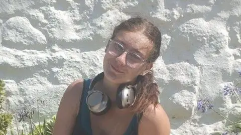 MYTIME Young Carers A girl with dark hair in a ponytail poses for a picture in front of a white stone wall. She is wearing glasses, smiling, and has a pair of headphones wrapped around her neck. 