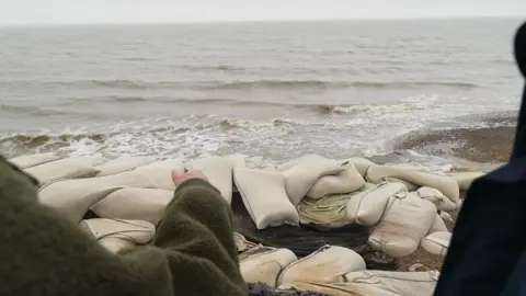 Martin Giles/BBC A view of the bottom of Hilary's garden looking down toward the sea and coastline. Sandbags that have been used a defence have been stacked on the beach. 
