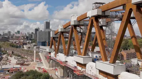 Network Rail An view of a railway viaduct under construction, with supportive structures and scaffolding in place around it. Tall city buildings can be seen in the distance on a cloudy day. 