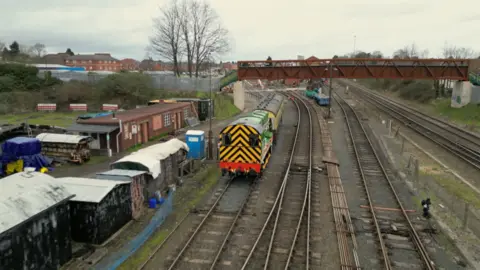 A green diesel shunter on a railway line heading under a footbridge. The engine has black and yellow diagonal stripes on the back and a red bumper. Alongside the track are a collection of sheds