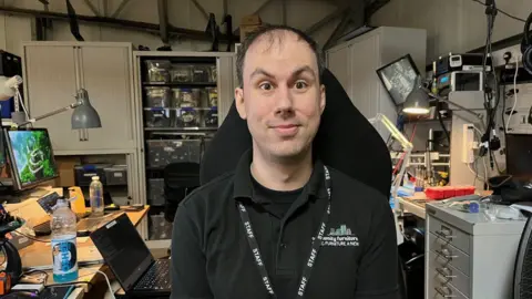 Tyler Potter looks at the camera as he sits in a room full of computers. He has short, brown hair and is wearing a black polo shirt and a black lanyard saying staff.