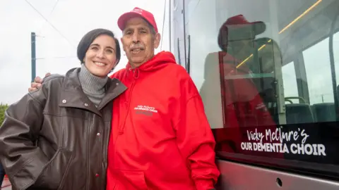 Vicky McClure and a man in a red 'Dementia Choir' jumper standing next to a tram with the words 'Vicky McClure's Our Dementia Choice' written on the side