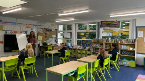 A classroom. It has five wooden tables with green table legs and green chairs. There are four students, facing away from the camera towards the front of the classroom. There is a female teacher at the front next to a whiteboard. 