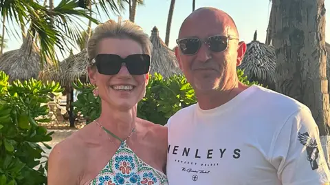 Jo Fuller/Brain Tumour Research/PA Wire Jo with her husband Wayne on holiday. Jo has short blonde her and is wearing a strappy summer dress with a tile print. Wayne, standing on her left, is wearing a white T-shirt which reads 'Henleys'. They are both smiling. 