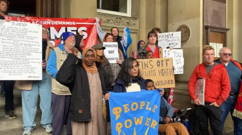 Several men and women, most holding posters, placards and messages, stand on the steps of the council house in protest at the plans being passed. The colourful messages say 'People Power' and 'Hands off Druids Heath'.
