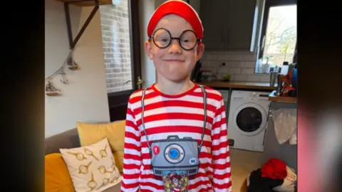 Rufus, wearing rounded glasses and a red striped shirt, standing for the camera with a kitchen in the background.