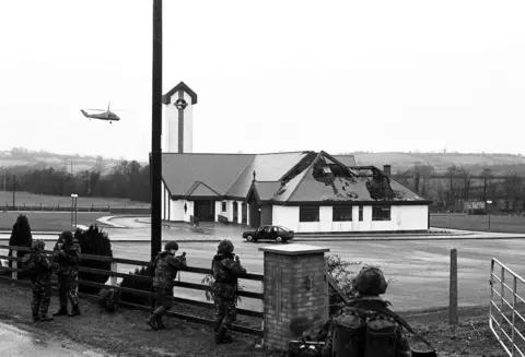 Pacemaker Five soldiers wearing camouflage gear stand guard beside the carpark of St Patrick's Church, which has a partially crumbling rook. A car sits stationary in the car park while a helicopter flies overhead. 