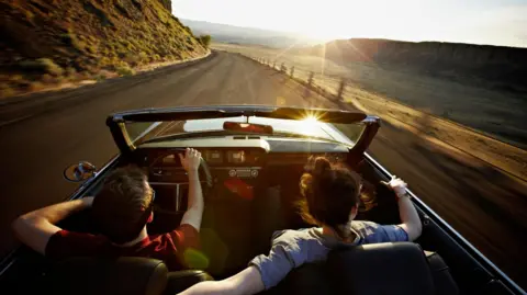 A young man and woman in a convertible car driving along a mountain road towards the sunset. She is driving and has her arm around the back of his seat.