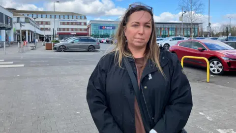 Henry Godfrey-Evans/BBC A woman with brown hair in a busy car park and lots of shops in the background.