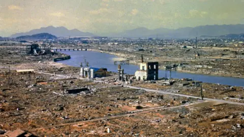 Getty Images The destruction caused by the nuclear explosion over Hiroshima. The landscape is largely flattened with visible debris from properties. The shells of some properties near a wide river as visible. The ground looks brown.