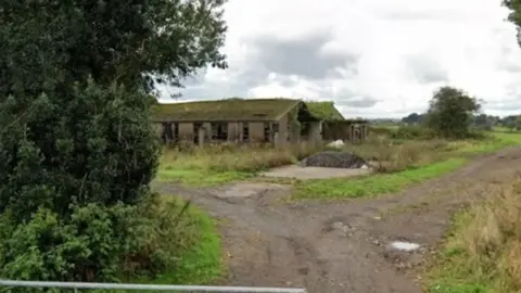 Google A single-storey building with its windows and doors missing and a green-covered roof at the end of a footpath with bushes in the foreground