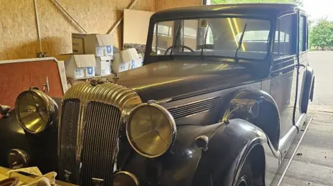 A vintage Daimler vehicle, in a storage unit and surrounded by cardboard boxes.