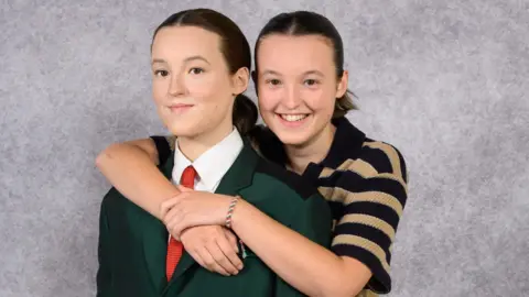 Madame Tussauds London Bella Ramsey wearing a black and cream hooped top stood behind a waxwork of them, with their arms around the waxwork's shoulders.