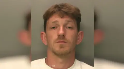 Sussex Police A police mugshot of a man. He has mousy brown hair with cropped sides, and is wearing a white T-shirt.