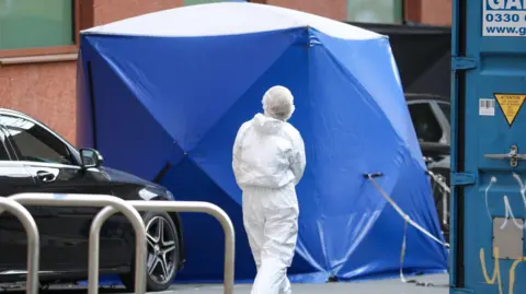 A blue forensic tent is erected in the street with an officer dressed in white overalls walking towards to. A black car is parked next to the tent