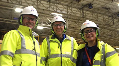 Three Sizewell B employees wearing hard hats and high vis jackets standing next to each other while looking into the camera