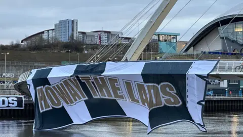 A large  achromatic  and achromatic  emblem  which reads 'Howay the Lads' hanging disconnected  the broadside  of the Millennium Bridge.