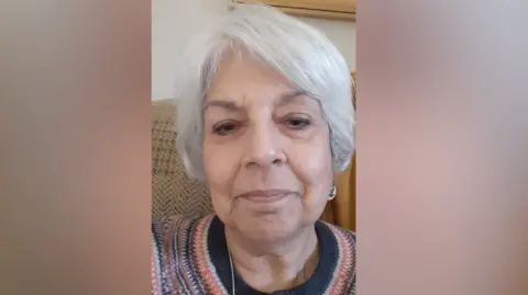 Satti Collins Satti Collins has white hair, a colourful patterned jumper and is sat on a sofa.