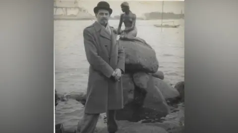 Robin Maudsley A black and white photo of Max standing at a harbour and wearing a long overcoat and a bowler hat. He has a moustache and is looking at the camera with a neutral expression on his face and his hands clasped together in front of him.