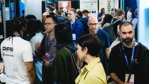 Other A crowd of people wearing lanyards in a big room attending the Develop:Brighton expo. A banner at the back says Indie Showcase, Vote Now.