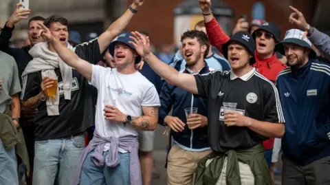 A group of Oasis fans in Cardiff with their arms in the air with some holding pints of beer. They are wearing the band's merchandise, including sport shirts with the logo on, along with bucket hats and are standing in a street. 