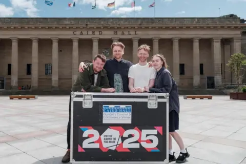 Grant Anderson Three young men and a woman stand outside a large hall in a city square. They are leaning on a large packing case with the words Scottish Album of the Year 2025.