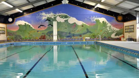 Shropshire Council A swimming pool with blue tiles. On the wall behind it is a large mural of a blue/green wave. Inside the wave is a large whale, schools of fish and dolphins.