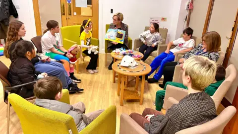 Martin Giles/BBC A group of children and adults sit around a table and look at a picture book. The room is brightly lit and the children look engaged. 