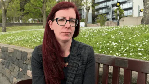 Kaye Nicholson is a woman with shoulder-length red hair and glasses. She is wearing a dark checked jacket. 