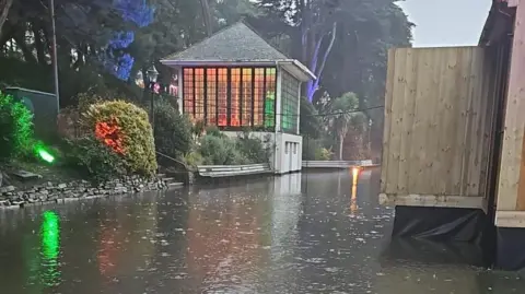 A flooded Bourne Stream with the bandstand and shrubs on the bank on the left. On the right is a raised wooden structure. The base is partly submerged.