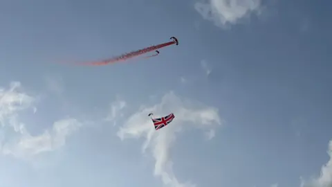 Three parachutes in the sky. Red smoke is coming from one of the red parachutes and a Union flag is being flown. A blue sky and clouds are behind the display team.