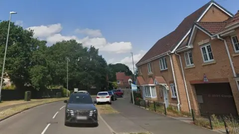 A row of houses on a residential road. There are three cars parked on a grass verge and two cars on the road. There are trees on the opposite side of the road.