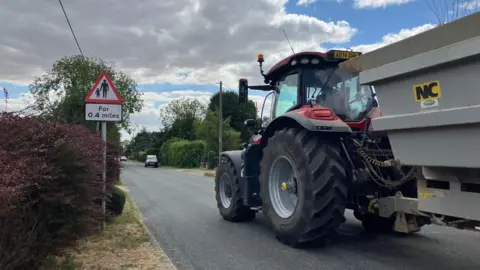 Owen Sennitt/BBC A pedestrian warning sign is on the side of a road, which is lined with shrubs and trees. A large tractor with a large trailer is travelling along the road, and there are other vehicles in the distance.