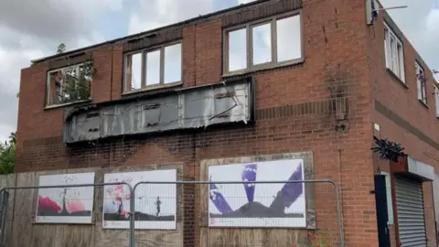 The exterior of an old red brick two-storey building. It has wooden boards with three faded posters on stuck on. There are metal railings in from of the wooden borders. There are three windows on the second floor of the building with a broken sign below. The sky is cloudy. 