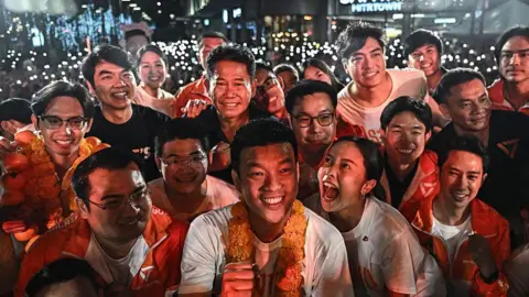 Getty Images Young Thai voters wearing white shirts and orange jackets, holding up their phones as lights at an election campaign rally