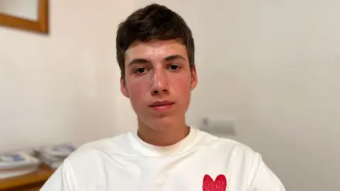 A teenage boy looks straight faced into the camera , posing for a picture, wearing a white t-shirt featuring a small red heart logo