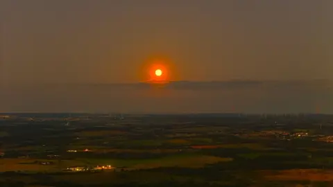 Kevin Hamill A bright orange sun glows in the distance over a dark landscape of fields. A small slither of cloud runs across the sky and a windfarm can be seen in the distance as well as shining roofs and windows of a town or village.