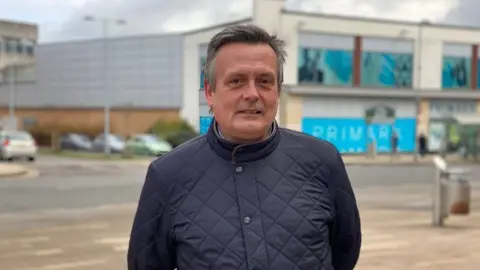 Dan Pickard with medium-length hair wearing a dark black padded jacket. He is standing on a pavement outside a two-storey Primark store with blue and white walls.