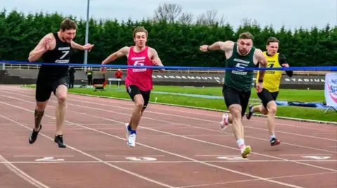 Bobby Gavin Runner wearing green vest and black running shorts, second from right, dips to break the tape on red athletics track just ahead of runner in black vest far left, one in red vest, second left, and another in yellow vest to his right.