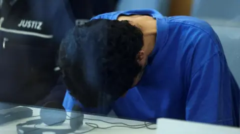 A Syrian national sits, his head bowed and wearing a blue T-shirt, behind a glss screen in a court room in Germany 