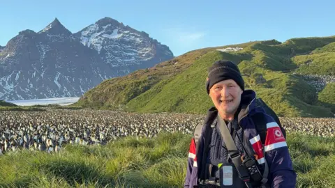 Ben Osborne Ben Osborne is wearing outdoor gear and carrying camera equipment stands in tall grass with a vast colony of penguins stretching across the landscape behind them, with mountains and a glacier in the background under clear skies.